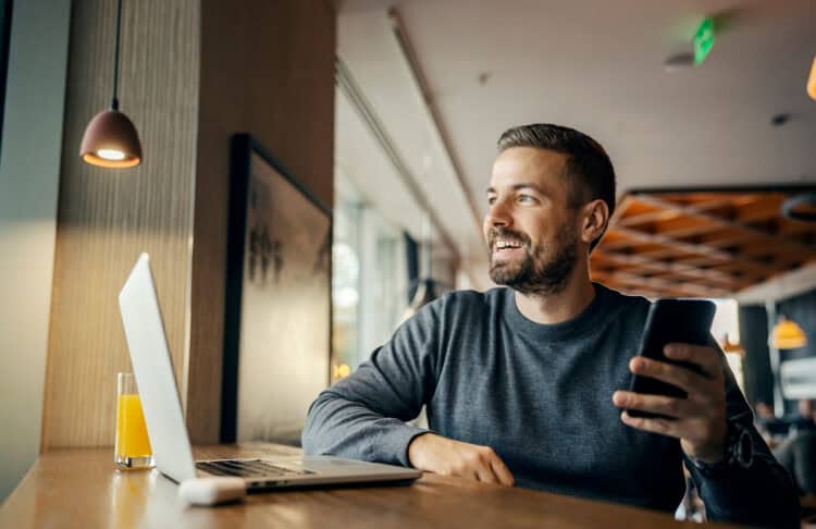 A happy freelancer is working on social media by using multiple technologies at coffee shop.